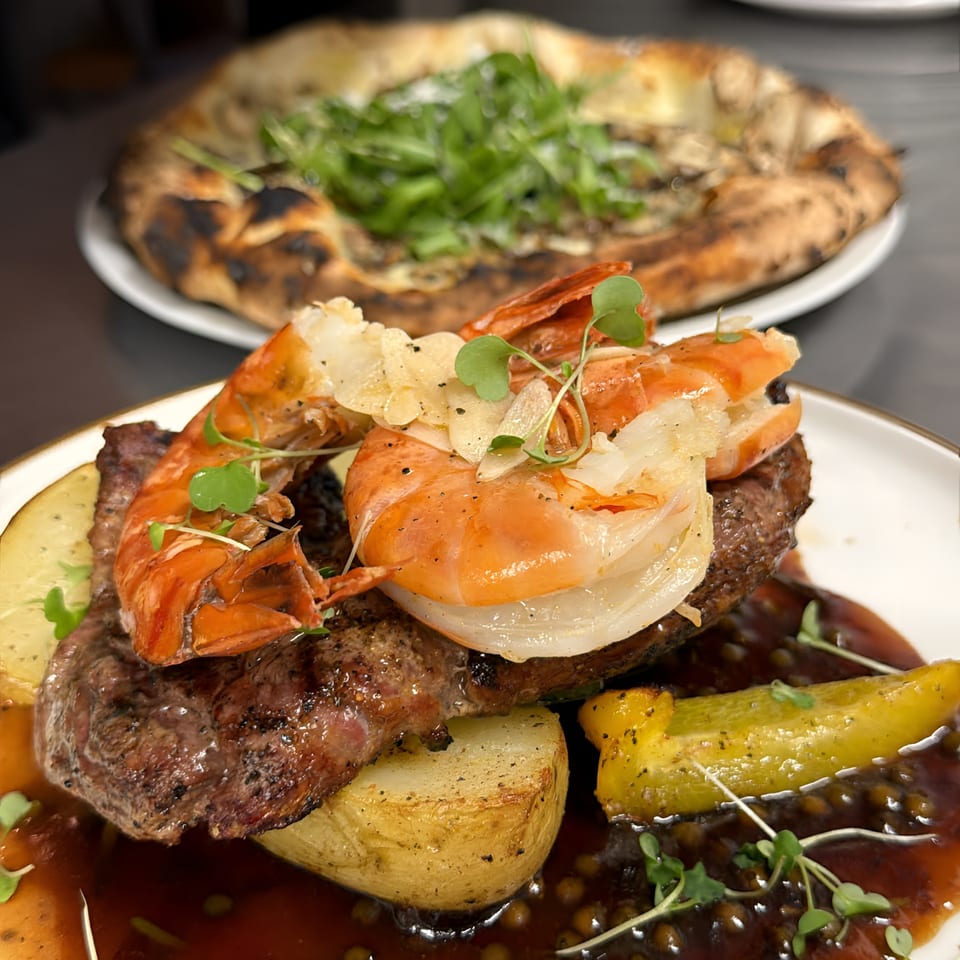 Grilled steak topped with lobster, roasted potatoes, vegetables, microgreens, and dark sauce, with a leafy pizza behind it.