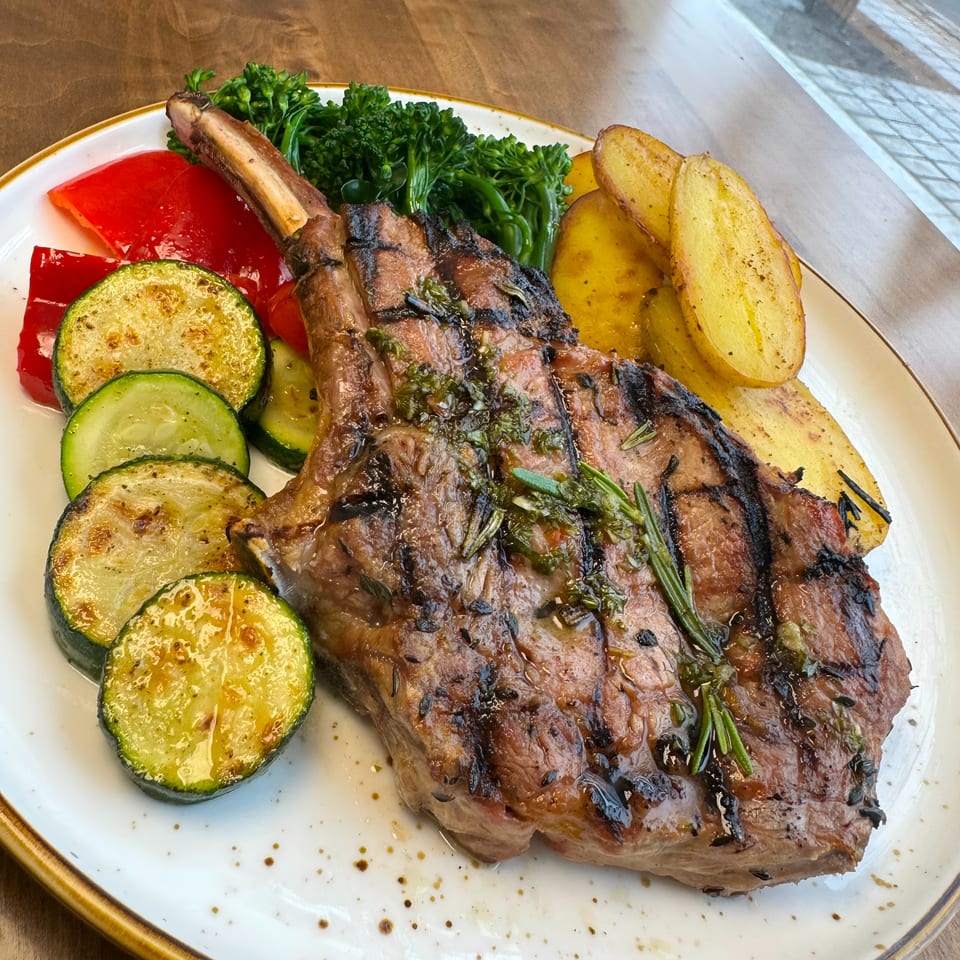 Grilled bone-in steak with rosemary, roasted potatoes, sautéed zucchini, red peppers, and greens on a plate.